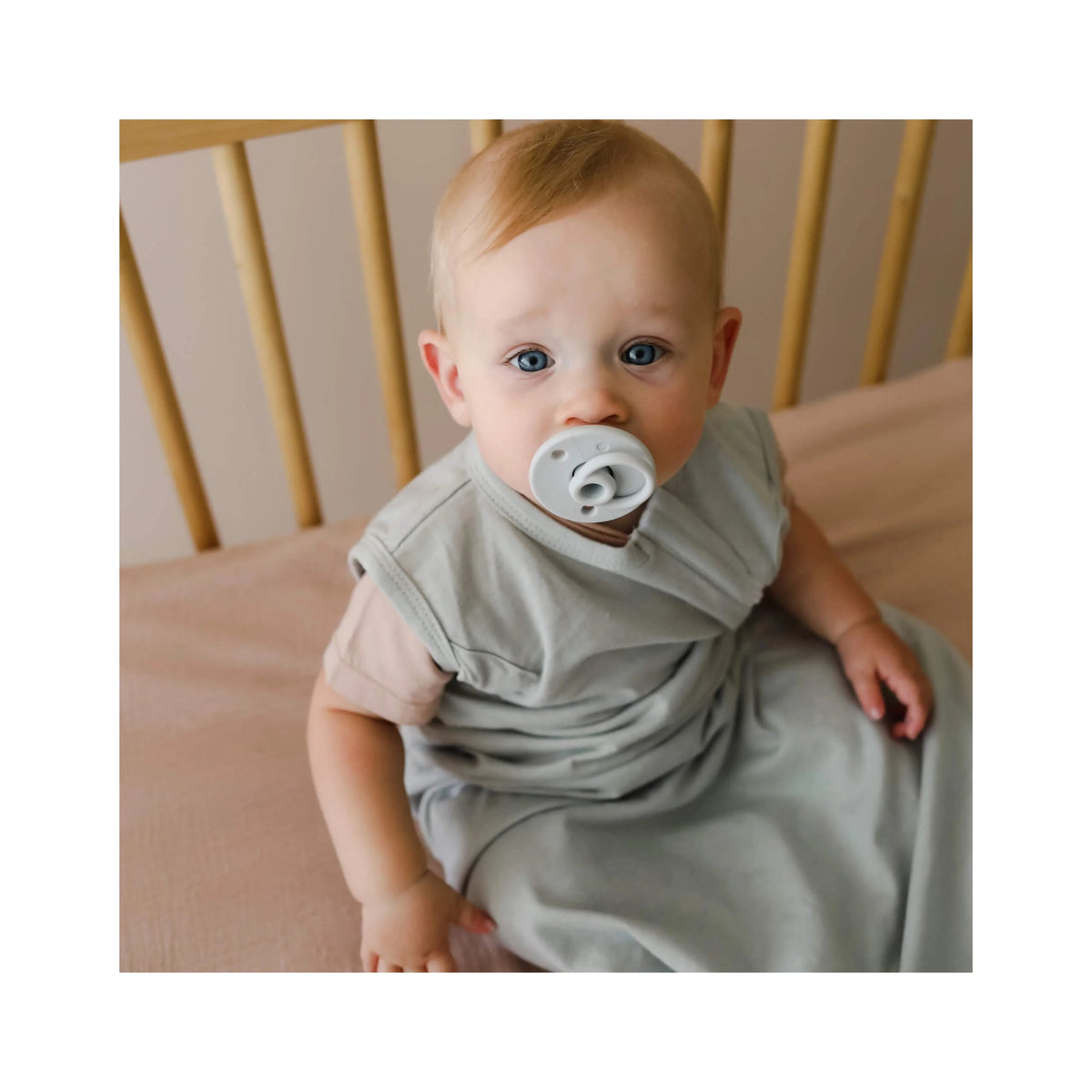 Baby sitting in a crib wearing a light gray outfit with a pacifier.
