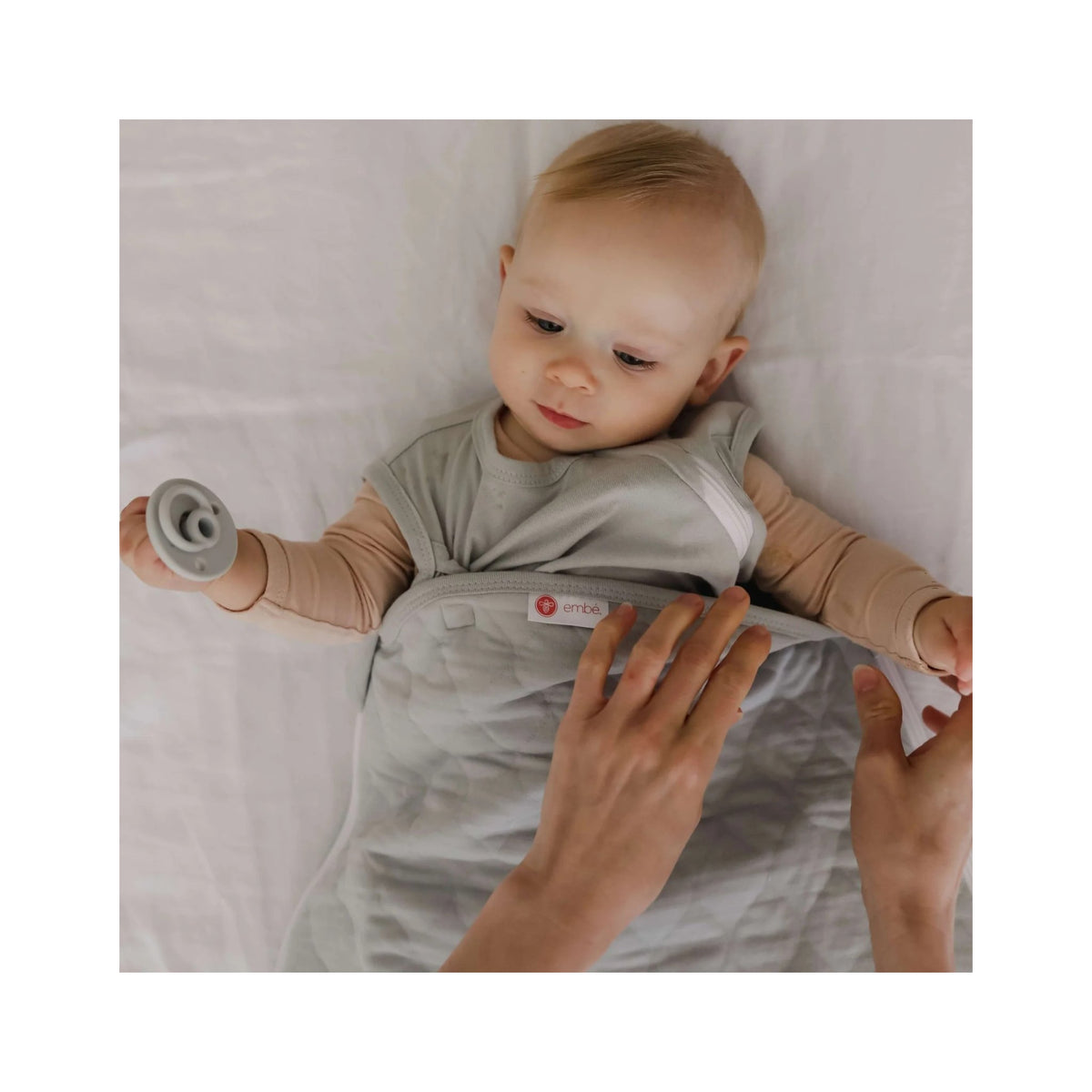 Baby lying on a white blanket with a person's hands holding a pacifier