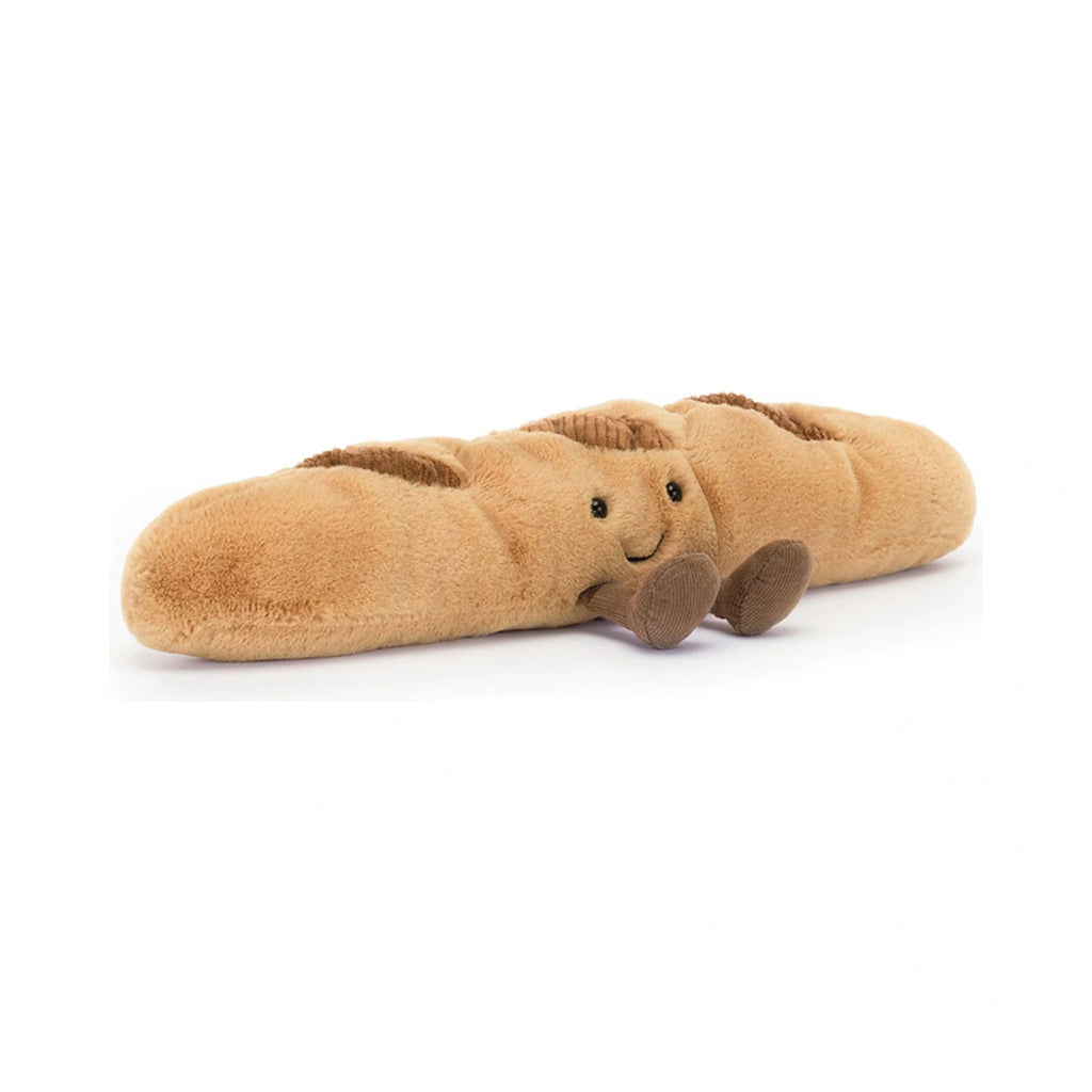 A golden brown Jellycat Amuseables Baguette plush toy features a smiling face, beaded eyes, textured crust details, and small brown corduroy feet lying flat on a plain white background.