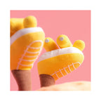 A close up detail of the Jellycat Amuseables Sun Roller Skates featuring soft yellow fabric boots with stitched white laces, white soles, and yellow wheels on brown ribbed legs against a solid pink background.