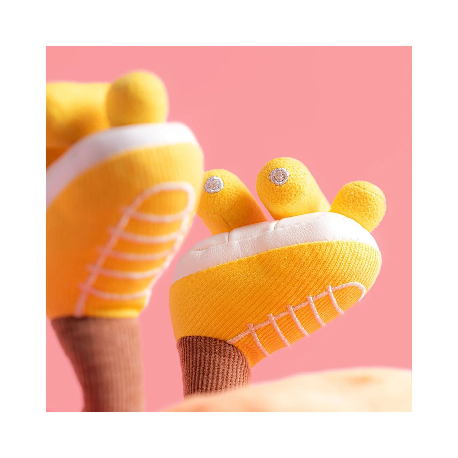 A close up detail of the Jellycat Amuseables Sun Roller Skates featuring soft yellow fabric boots with stitched white laces, white soles, and yellow wheels on brown ribbed legs against a solid pink background.