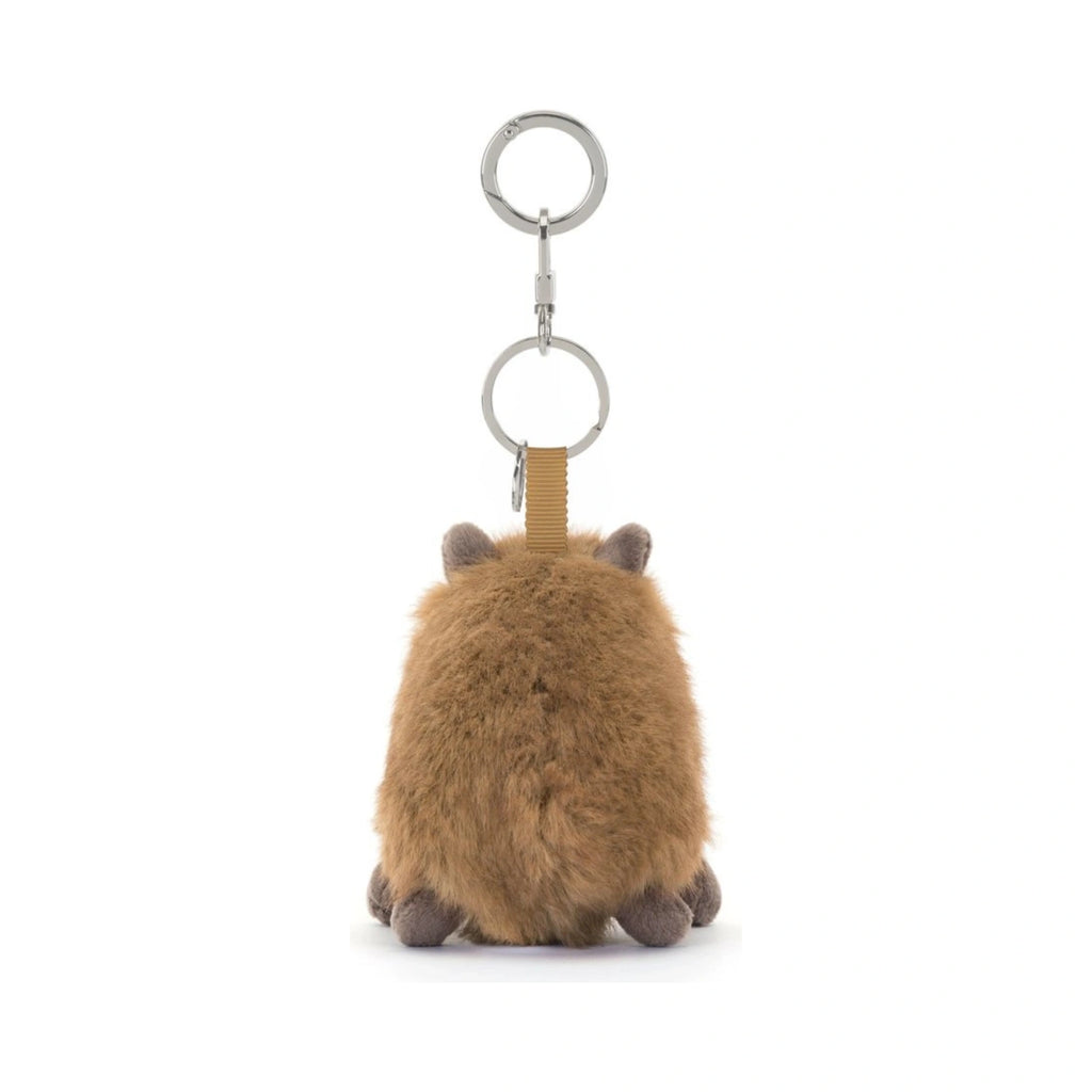 A back view of the Jellycat Clyde Capybara Clip On Charm, featuring fluffy brown plush fur, tiny ears, and small feet, suspended from a silver metal key chain clasp by a tan textured fabric loop.