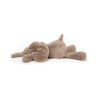 The Jellycat Smudge Elephant Medium plush toy is depicted lying flat on its stomach, showcasing its ultra soft taupe fur, oversized floppy ears, and relaxed pose.