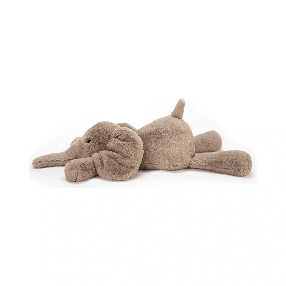 The Jellycat Smudge Elephant Medium plush toy is depicted lying flat on its stomach, showcasing its ultra soft taupe fur, oversized floppy ears, and relaxed pose.