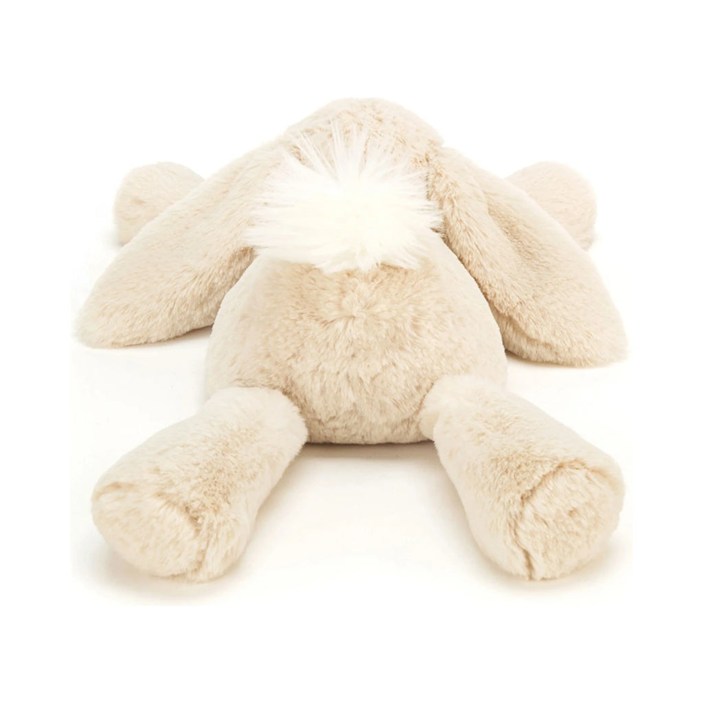 The back view of a beige Jellycat Smudge Rabbit Medium plush toy lying flat, featuring long floppy ears and a fluffy white tail.