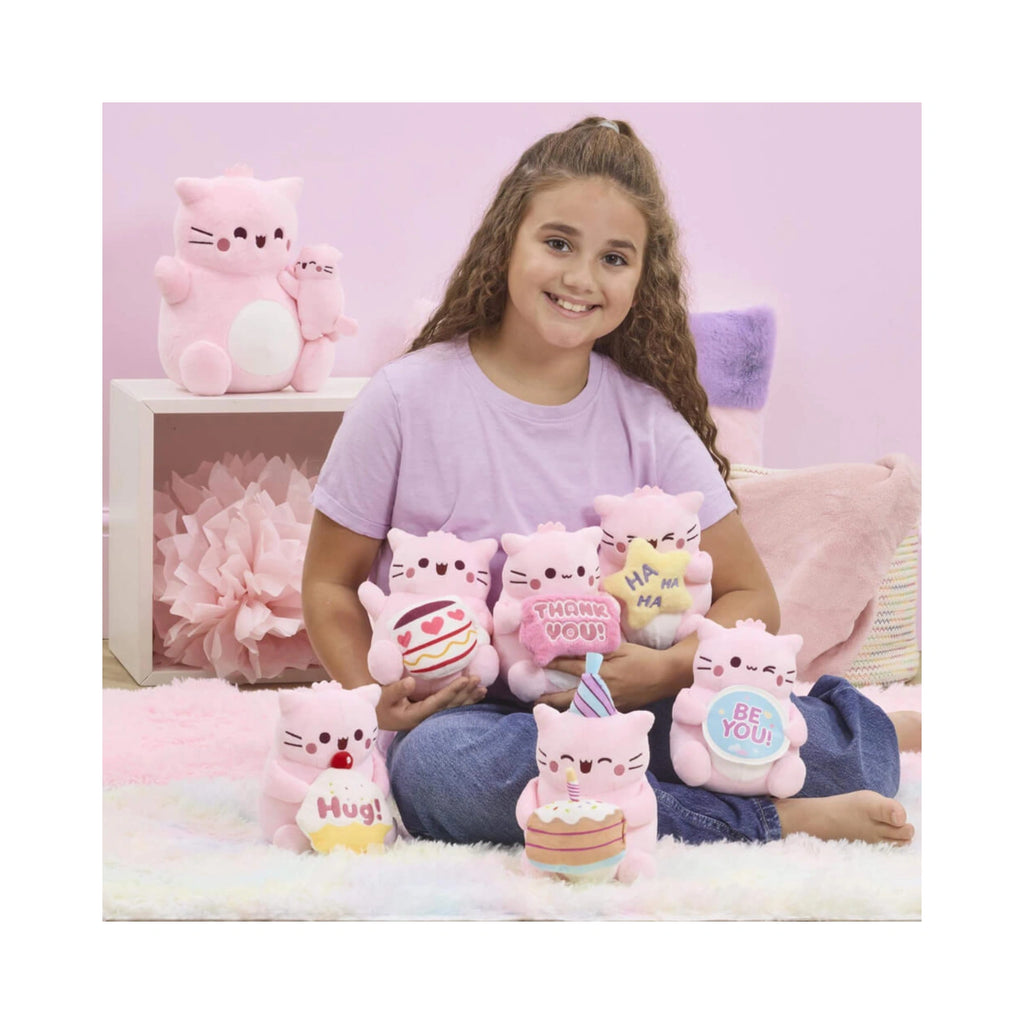 A young girl sits on a fluffy white rug holding a collection of Pembe the Pink Cat small soft toys, including one holding a blue circular sign that says Be You and others with cakes and greeting signs.