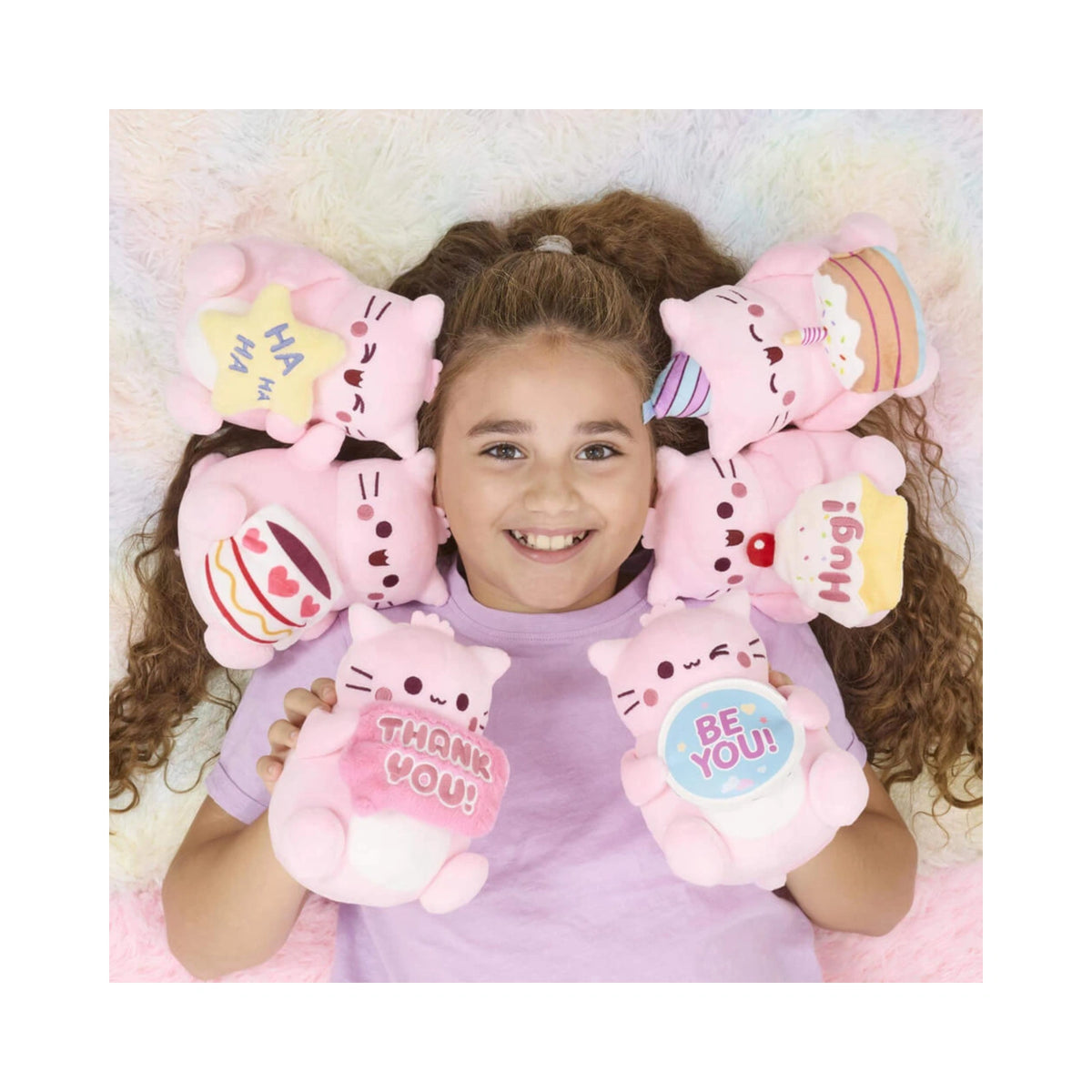 A young girl smiles while lying down surrounded by Pembe the Pink Cat soft toys, including the PEMBE BE YOU SMALL SOFT TOY and other themed pink plushies with colorful signs and cupcake designs.